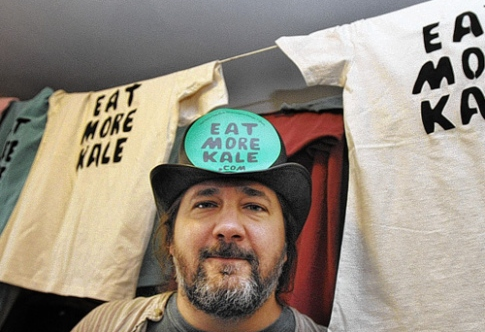 Bo Muller-Moore wears a hat that reads Eat More Kale .com, in front of shirts with the same call to action