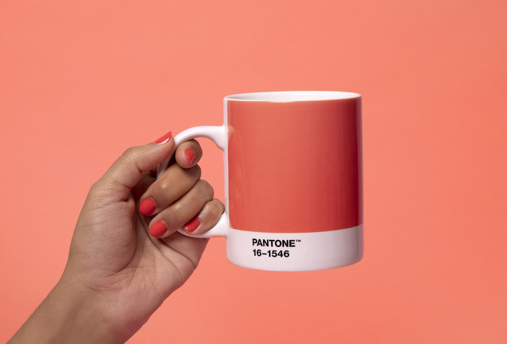 A hand holds the handle of a mug with the Pantone color of the year on it - the color is coral pink